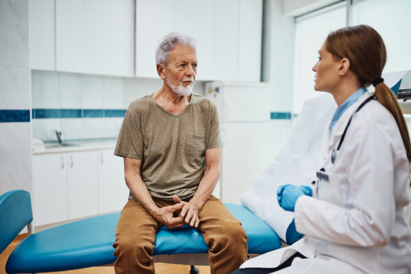senior-male-patient-consulting-doctor-exam-room A senior man sitting on a blue examination table in a medical clinic, discussing his health with a female doctor wearing a white coat and blue gloves.